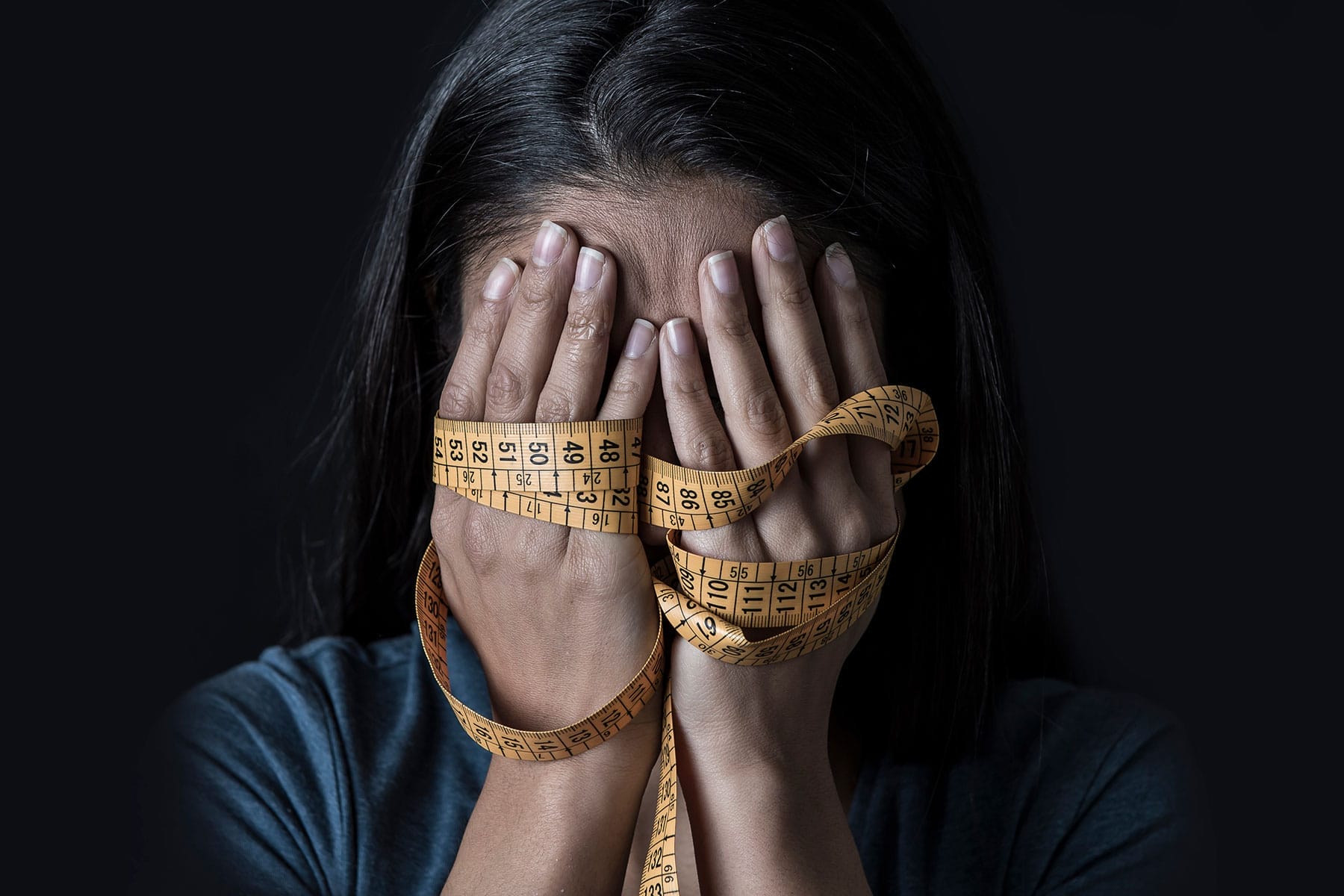 Woman hides face in hands and measuring tape as she wonders, "What is Bulimia?"